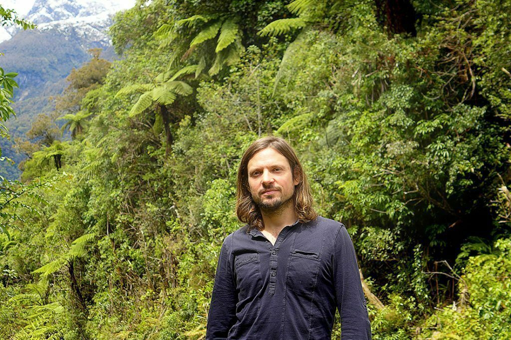 Lush jungle and a snowy peak in the back. Vincent in the front wearing a black buttoned-shirt looking strong and confident.