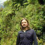 Lush jungle and a snowy peak in the back. Vincent in the front wearing a black buttoned-shirt looking strong and confident.