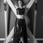 Black and white shot of Electra standing in front of a big wooden frame that has some straps for tying people.