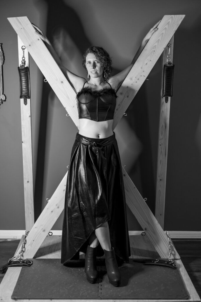 Black and white shot of Electra standing in front of a big wooden frame that has some straps for tying people.