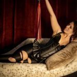 Lying down on a sofa. Goddex Electra in leather outfit holding a red flogger.