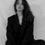 Black/white shot of calypso in dark formal clothing. Sitting on a chair looking professional and serious.