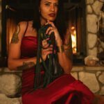 Cozy room with a lit fireplace. Lilith sitting in front of it in a velvety red dress.