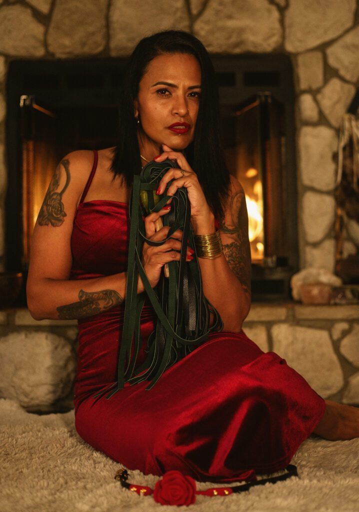 Cozy room with a lit fireplace. Lilith sitting in front of it in a velvety red dress.