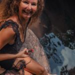 Sitting on a rock with a big smile holding a red flower in her hands.