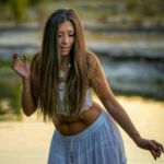Golden hour. Stephanie standing in water with her eyes closed and loose hair hanging down to the front of her body.