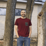 Adam standing next to trees with his hands in the pockets of his jeans.