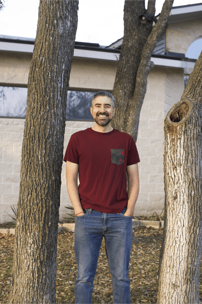 Adam standing next to trees with his hands in the pockets of his jeans.