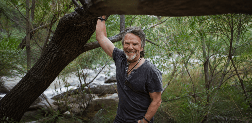 Rich by a river holding his hand to a big tree.