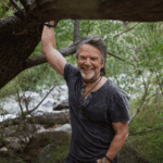 Rich by a river holding his hand to a big tree.