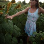 Passed flowering sunflower field and Liv touching the leaves in a light-blue dress with flowers on it.