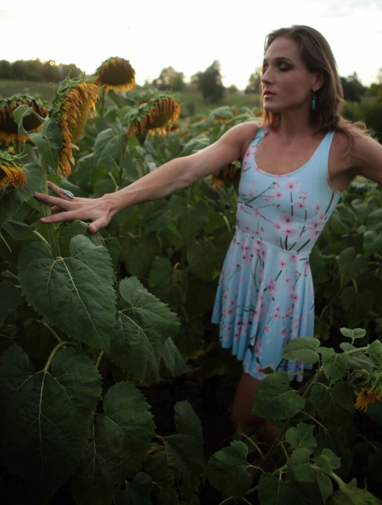 Passed flowering sunflower field and Liv touching the leaves in a light-blue dress with flowers on it.