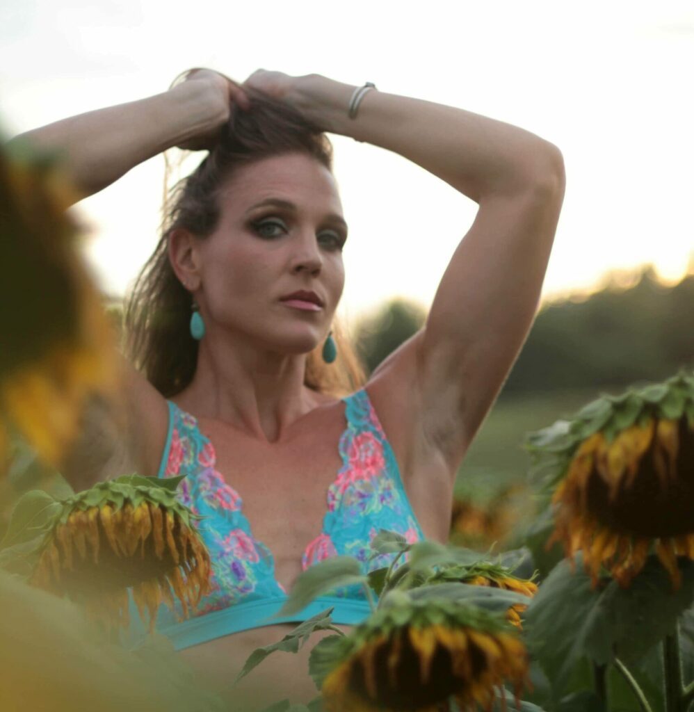 Tantra & Intimacy Coach standing in a field of sunflowers in a blue swimsuit-like top.