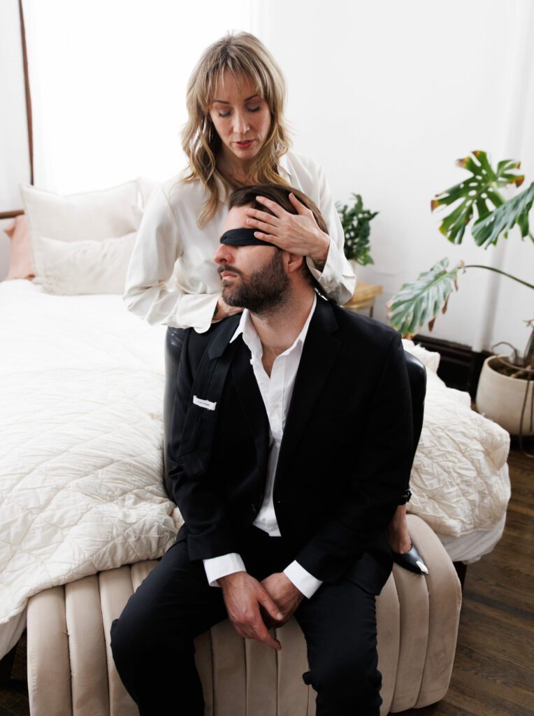Practitioner standing in front of a bed. A man in black formal clothing, wearing a blindfold is sitting in front of Butterfly.