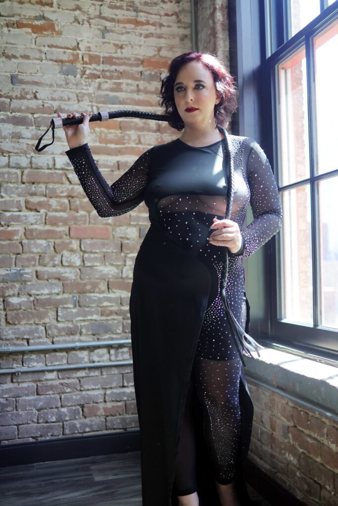 Tigress Lily wearing black and holding a flogger.
