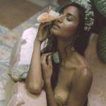 Intiacy Coach naked upper body with seashells covering nipples holding flowers against her face while eyes are closed.