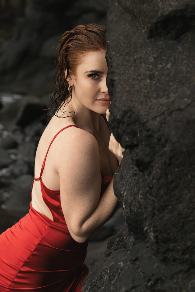 Jo standing by a rocky natural wall wearing a red velvety dress.