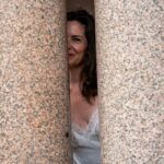 Mistress Aviva peeking out from between two stone pillars.