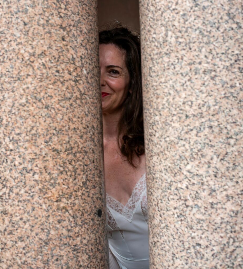 Mistress Aviva peeking out from between two stone pillars.