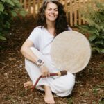 Aiua sitting outside on a mulchy ground holding a shamanic drum.