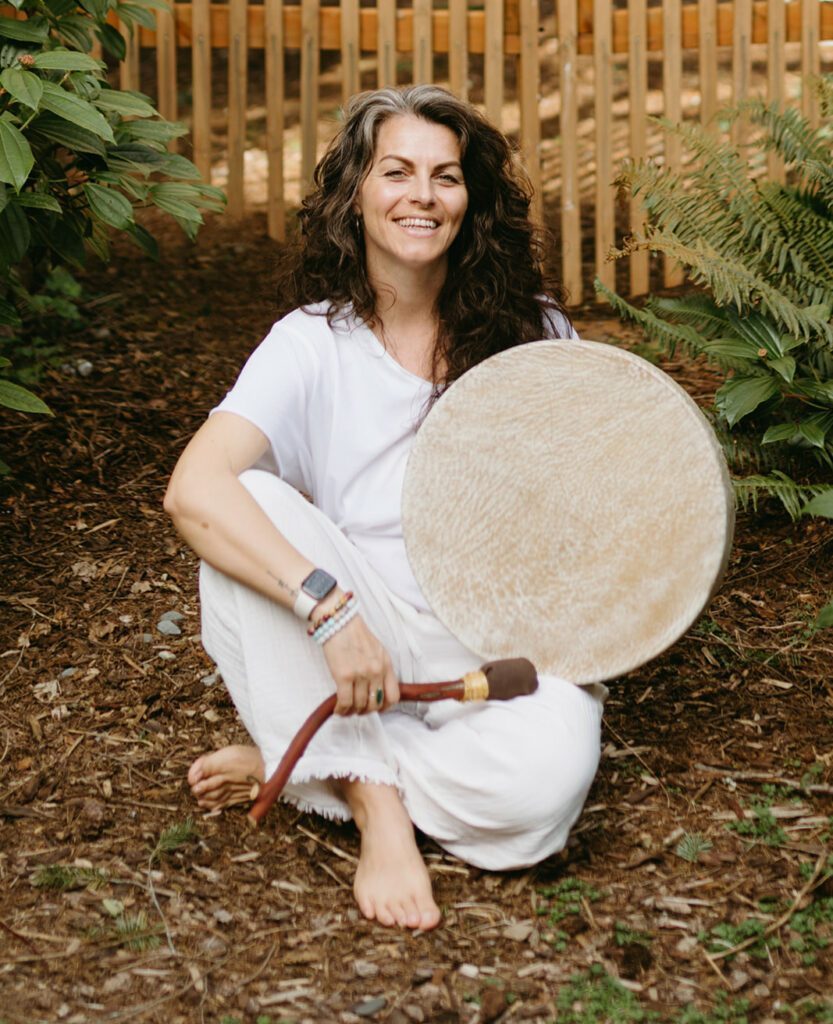 Aiua sitting outside on a mulchy ground holding a shamanic drum.