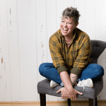 Kel sitting cross-legged on a chair and laughing.