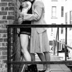 Amada on a rooftop balcony embracing a male client with a bag over his head from the back.