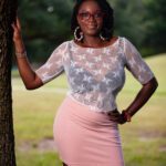 Standing next to a tree wearing a pink skirt and white trasparent silky top with black bra showing underneath.