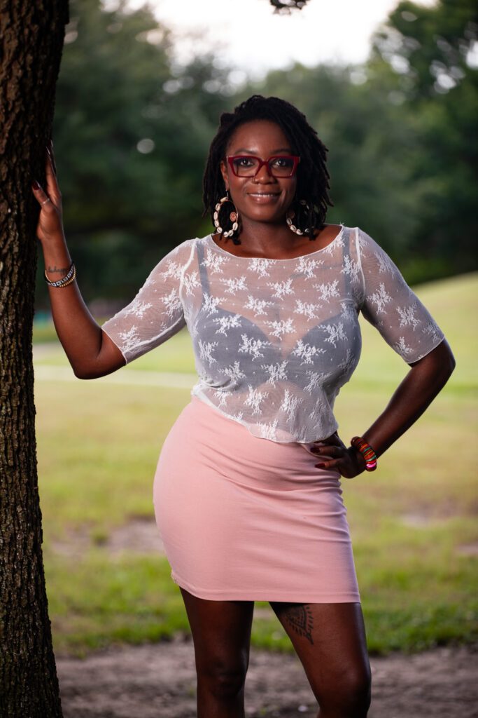 Standing next to a tree wearing a pink skirt and white trasparent silky top with black bra showing underneath.