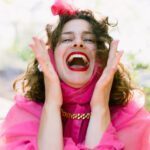 Allie in pink fluffy dress holding hands by her head with a big open smile.