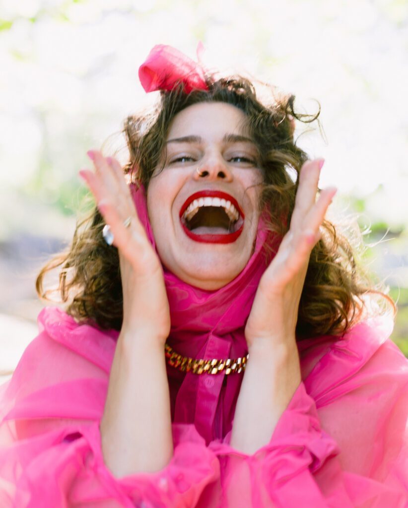 Allie in pink fluffy dress holding hands by her head with a big open smile.