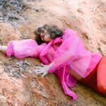 Lying down in the nature on a brown sandy surface with some dry leaves. Wearing pink and red.