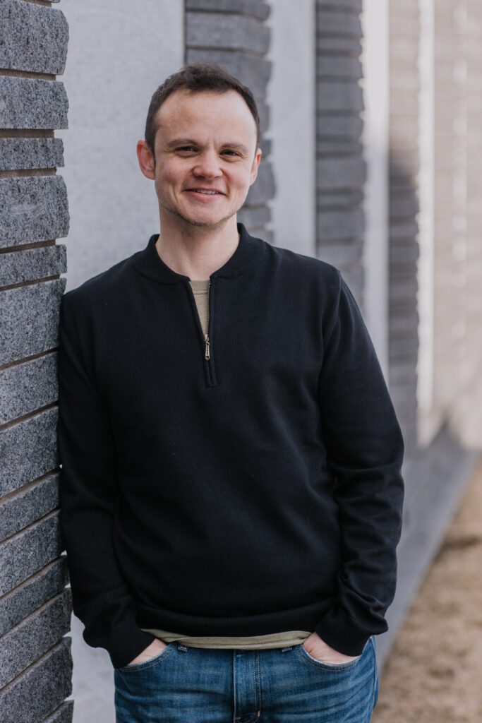 Grey leaning on a wall and smiling.