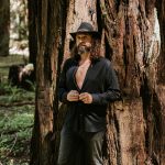 Daniel in a black outfit and grey top hat in front of a big redwood.
