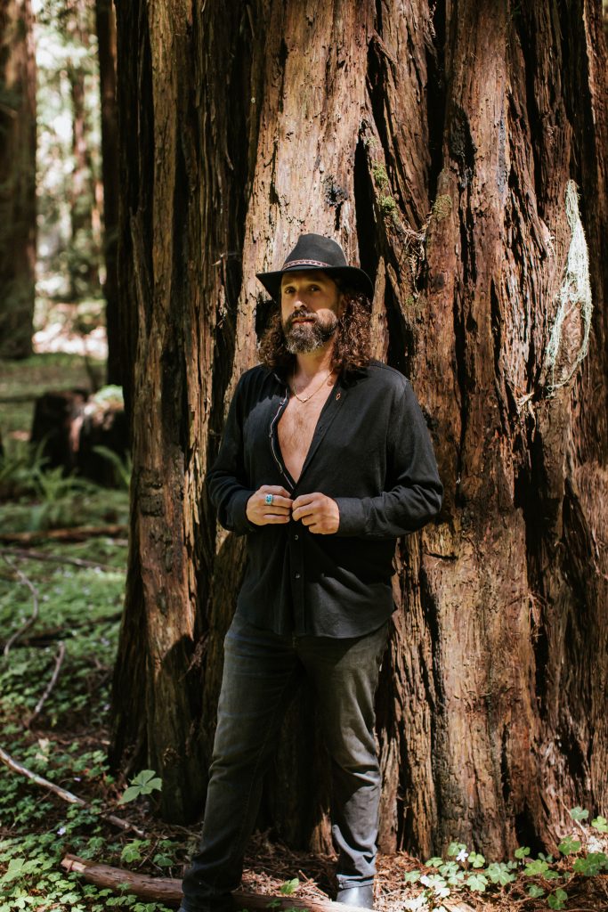 Daniel in a black outfit and grey top hat in front of a big redwood.