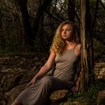 Gina Marie in a long grey dress leaning against a tree.